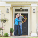 A happy senior couple standing together outside their home, at the front door, smiling and waving.